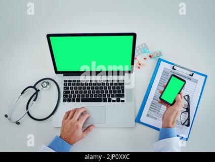 Technology helps keep track of it all. High angle shot of an unrecognizable doctor using a laptop and cellphone with a green screen in his office. Stock Photo