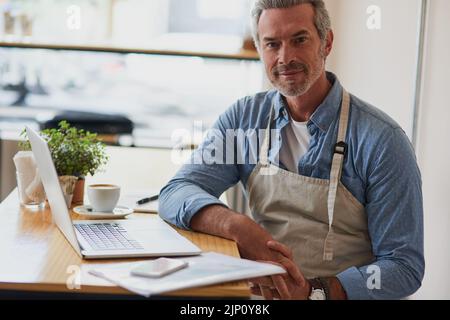 I found success in coffee. Portrait of a handsome mature man doing some paperwork and using a laptop in his cafe. Stock Photo