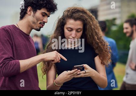 Yes thats how you spell my name. two cheerful young friends browsing on a cellphone together while standing in a park outside during the day. Stock Photo