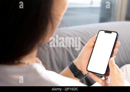 Holding smart phone, young woman sitting on sofa and holding smart phone with mockup white blank display. Vertical empty screen for social media apps. Stock Photo