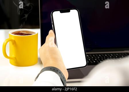 Holding smartphone, woman hand holding smartphone white blank screen for mock up. Caucasian female using laptop and looking to phone empty display. Stock Photo