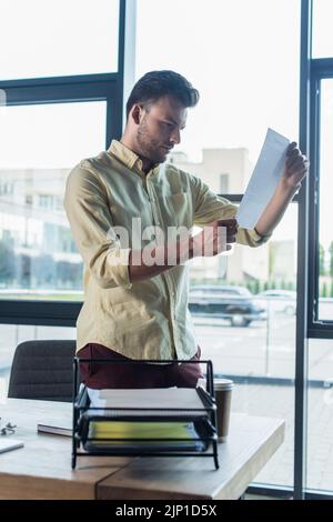 Businessman looking at document near coffee to go and papers in office,stock image Stock Photo