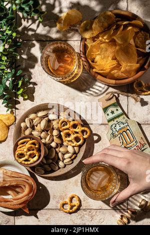 Beer outdoor party concept. Woman's hand takes a glass of beer from the table with different snacks. Still life on tile background with hard shadows, Stock Photo