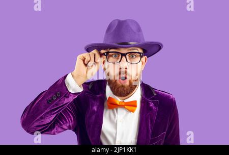Stylish extravagant man looks at you with surprised and shocked expression on purple background. Stock Photo