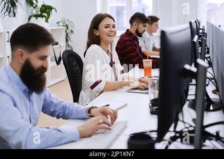 Junior female professional working in modern IT office. Group of young and experienced programmers and software developers sitting at desks working on Stock Photo