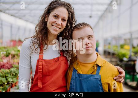 Experienced woman florist posing with young colleague with Down syndrome in garden centre. Stock Photo