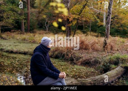 Young man in warm clothes sitting on a fallen tree near river, looking at the forest and enjoying the moment. Feeling harmony, reunion with nature in Stock Photo
