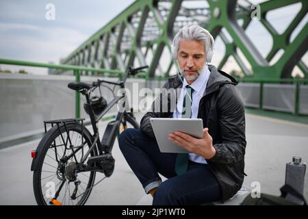 Businessman with bike sitting on bridge, using tablet and listening to music. Commuting and alternative transport concept Stock Photo