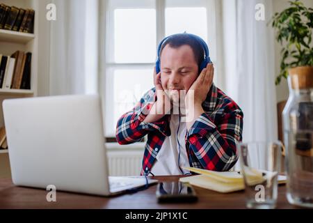 Young man with Down syndrome sitting at desk in office and using laptop, listening to music from headphones. Stock Photo