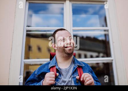 Portrait of happy young man with Down sydrome with backpack in street, smiling. Stock Photo