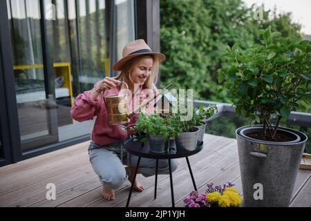 Young woman taking care of her herbs, watering them on terrace in tiny house, sustainable living. Stock Photo