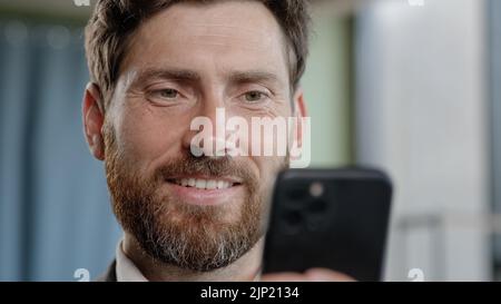 Close up head shot smiling bearded adult 40s businessman holding smartphone in office smile reading good news using online app middle-aged manager Stock Photo