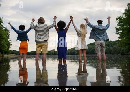 Multiracial group of young friends holding hands and standing in lake in summer. Rear view. Stock Photo