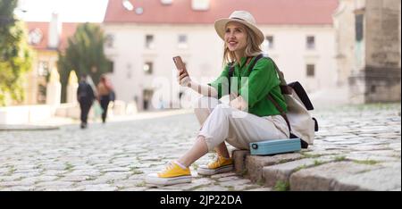Young woman travel alone in old city centre, sitting and using smartphone. Stock Photo