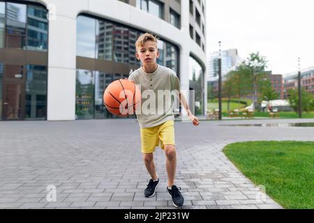 Caucasian boy driblling with basketball ball in public city park, looking at camera. Stock Photo