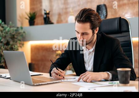 Concentrated clever caucasian business man, IT specialist, ceo, sits at the workplace in the office, works in a laptop, works on a new project, develop new app, strategy, study documents, graphs Stock Photo