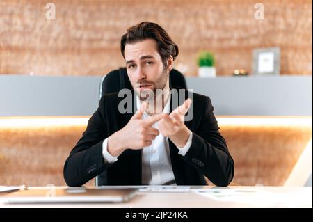 Serious elegante caucasian businessman, financial mentor, sits at a desk in a modern office, talking on webcam, having video call with client or colleague, recording online training course, explain Stock Photo