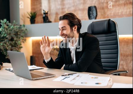 Online business meeting. Friendly attractive successful caucasian business man, top manager, in a suit, communicates with colleagues or clients via video communication, waves his hand, smiles Stock Photo