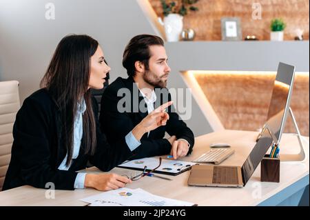 Two focused successful caucasian employees of the company, work together on a project while sitting at a table in a modern office, study financial charts, share information and ideas with each other Stock Photo
