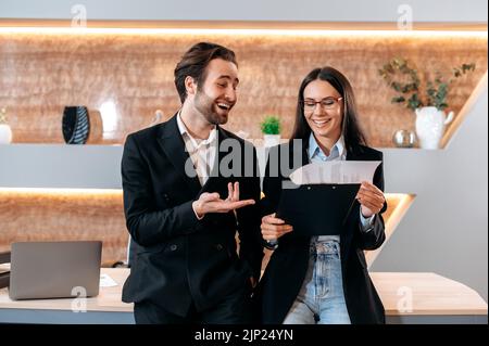 Positive successful confident business partners, caucasian man and woman, dressed in formal clothes, stand in a modern office near the desktop, discussing and sharing ideas, smiling friendly Stock Photo