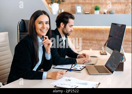 Positive pretty caucasian brunette young woman in a formal suit, financial manager, looks at the camera and smiles friendly while working. Successful coworkers working together in a modern office Stock Photo