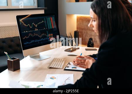 Successful cryptocurrency traders, caucasian man and woman, sit at a table in a modern office, looking at computer screen, work with stock charts and documents, analyze the growth dynamics Stock Photo