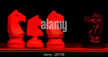 Black knight is standing on a chessboard against three white knights. Army of one. Selective focus. Neon red light in front of composition. Stock Photo