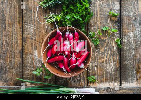 Long breakfast radishes in ceramic bowl and fresh springtime herbs on wooden board viewed from above. Organic homegrown vegetables, parsley, spring on Stock Photo
