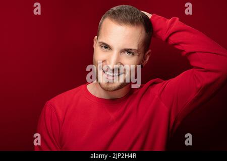 A Caucasian guy smiling at the camera with a joyful face and hand up high to his head moving shy but confident on a red background Stock Photo