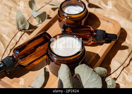 anti-aging collagen facial serum in dark glass bottle and face cream on wooden tray on craft paper background with eucalyptus branch. Natural Organic Stock Photo