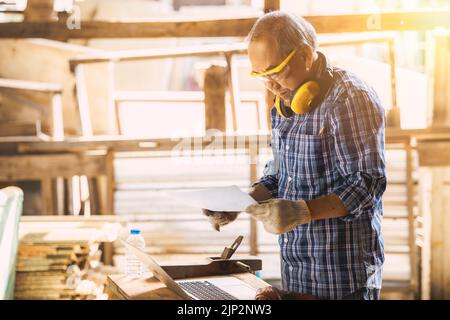professional senior carpenter using laptop computer for wooden furniture handcraft project working in wood workshop Stock Photo
