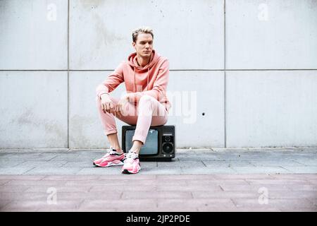 A handsome young Caucasian man in a pink outfit sitting on an old TV against a gray  wall Stock Photo