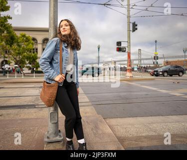 Unposed Portrait of a Beautiful Young Woman Waiting to Cross a Busy Downtown Street | Denim Jacket | San Francisco Wharf Stock Photo