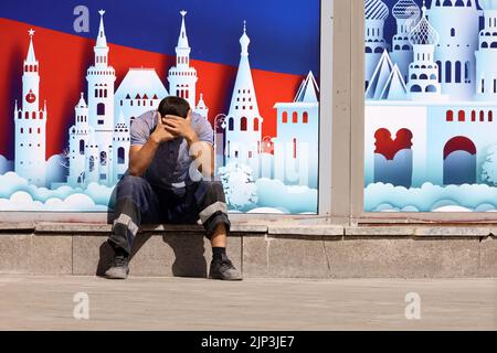 Man worker in uniform sitting on city street on background of decoration with Moscow landscape. Concept of unskilled labor in Russia Stock Photo