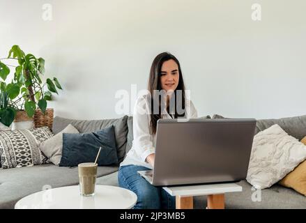 A happy businesswoman sitting on the sofa at home, using a laptop Stock Photo