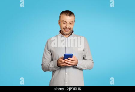 Mature Male Using Phone Texting And Websurfing Over Blue Background Stock Photo