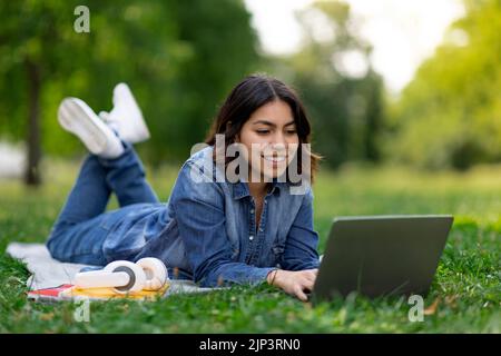 Smiling arab female student using laptop while lying on lawn outdoors Stock Photo