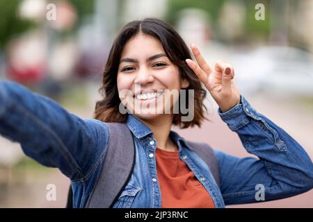 Cheerful Young Arab Woman Taking Selfie Outdoors, Showing Peace Gesture At Camera Stock Photo