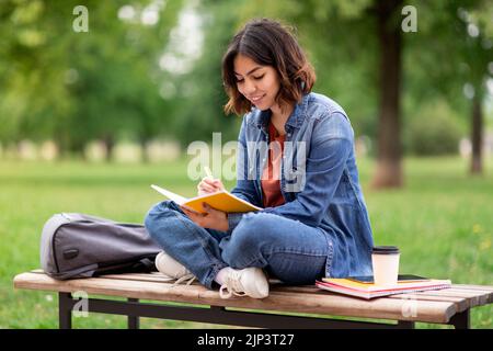 Arab female student writing in notebook while sitting on bench in park Stock Photo