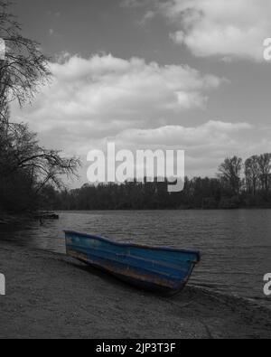 A vertical grayscale shot of a blue boat on the sand beach with a lake and a forest in front of it Stock Photo