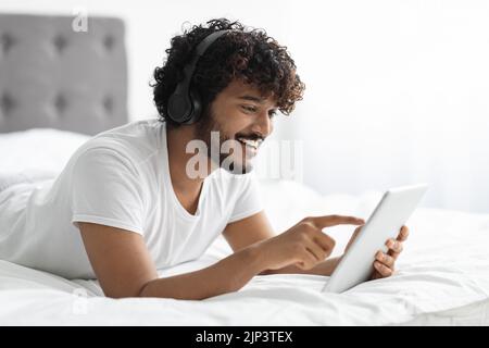 Relaxed hindu guy using digital tablet and headphones in bed Stock Photo