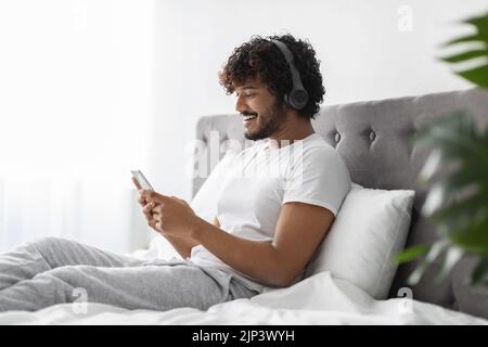 Cheerful hindu guy using cell phone and headphones in bed Stock Photo