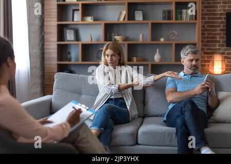 Upset angry european wife swears at husband with smartphone in office clinic interior. Problems in marriage Stock Photo
