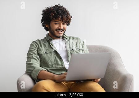 Smiling young indian guy sitting in armchair, chilling with laptop Stock Photo