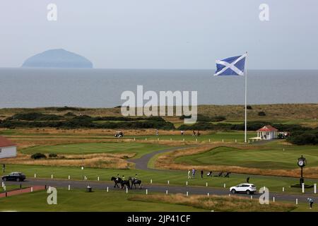 Trump Turnberry, Ayrshire, Scotland  UK..One of the largest saltire flags in Scotland  dominates the area and can be seen from miles around. This view seen from the Turnberry hotel with the starters hut for the first tee, practice greens and the 18th green. Also in the photo is the iconic large clock which bear Donald Trump's name. Golfers practice on the greens, with some teeing of from the 1st tee. Horse riders pass by On the horizon is the Firth of Clyde on the West Coast of Scotland with the iconic shape of Ailsa Craig Stock Photo