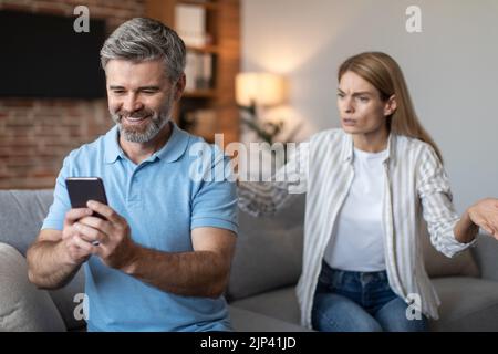 Angry upset adult european wife yelling at smiling husband with smartphone in living room interior Stock Photo