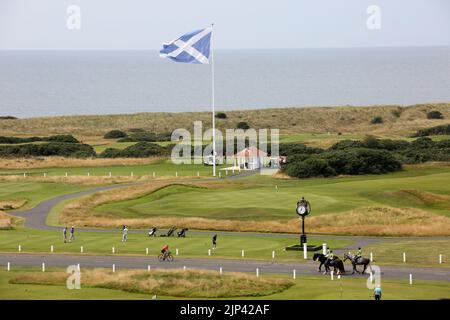 Trump Turnberry, Ayrshire, Scotland  UK..One of the largest saltire flags in Scotland  dominates the area and can be seen from miles around. This view seen from the Turnberry hotel with the starters hut for the first tee, practice greens and the 18th green. Also in the photo is the iconic large clock which bear Donald Trump's name. Golfers practice on the greens, with some teeing of from the 1st tee. Horse riders pass by On the horizon is the Firth of Clyde on the West Coast of Scotland Stock Photo