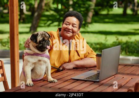 Portrait of smiling black woman using laptop in outdoor cafe with cute pug dog scene lit by sunlight Stock Photo
