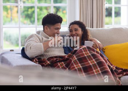 Happy biracial couple sitting on couch under blanket, drinking coffee and laughing in living room Stock Photo