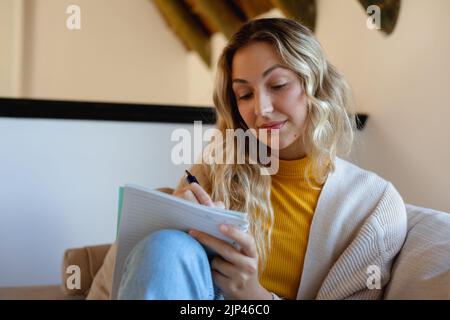 Happy caucasian woman sitting on sofa in living room, taking notes Stock Photo